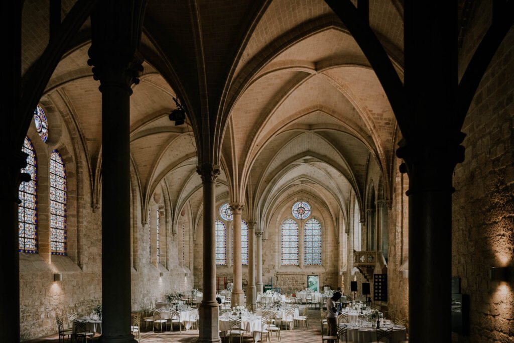 Salle de réception de l'Abbaye de Royaumont avec ses voûtes gothiques imposantes, ses vitraux colorés et son aménagement raffiné pour un dîner de mariage.