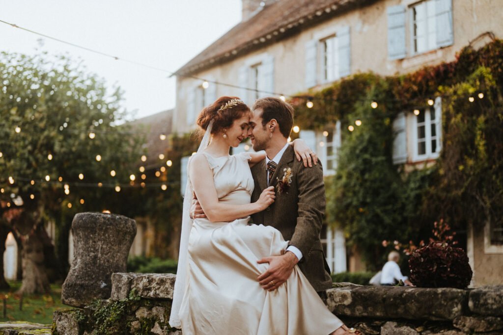 Un couple de mariés enlacés devant la façade romantique du Moulin de Launoy, illuminée par des guirlandes lumineuses en Île-de-France.