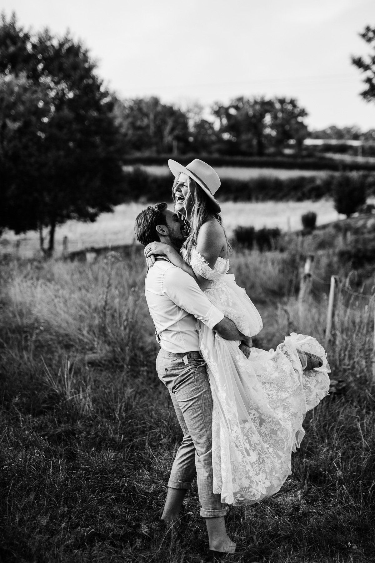 Black and white photograph of a laughing bride lifted by the groom in the French countryside.
